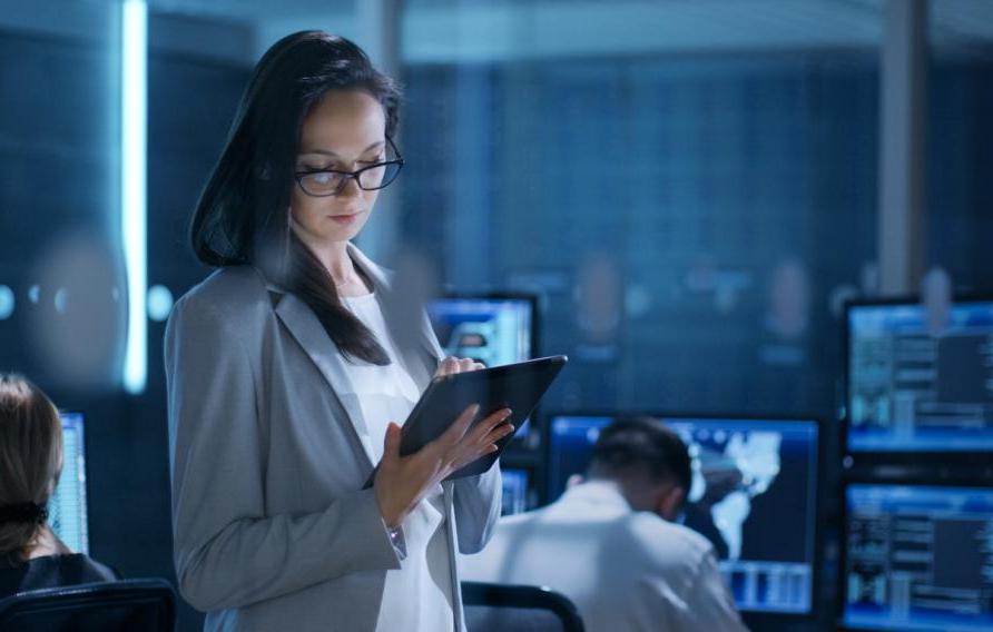 Young Female Engineer Uses Tablet in System Control Center. In the Background Her Coworkers are at Their Workspaces with many Displays Showing Valuable Data.